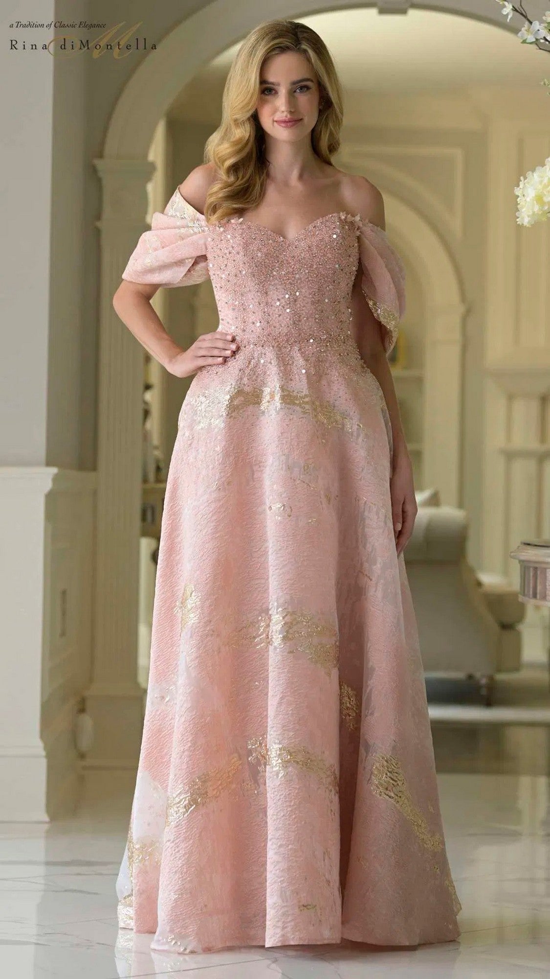 A woman stands indoors smiling with one hand at her side, wearing the blush Rina Di Montella RD3405 long formal dress—an organza, off-shoulder gown with a beaded bodice and gold accents.